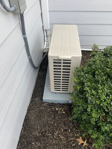 Outdoor air conditioner unit beside a house wall and shrubs on a concrete pad