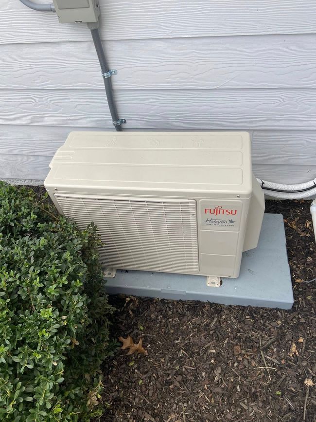 Beige outdoor HVAC unit beside a white house wall on a concrete pad near shrubs.