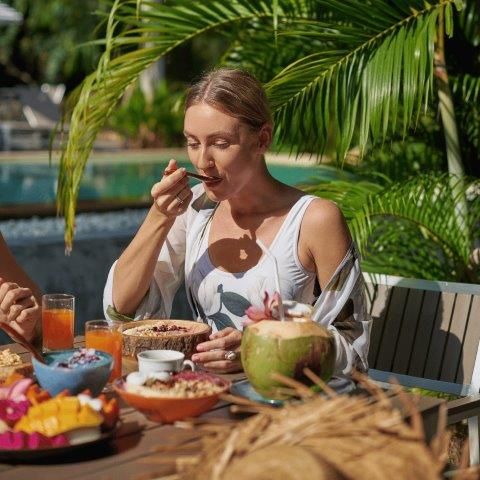 Woman eating breakfast outdoors by a pool, with tropical fruits and coconut, sunny day.