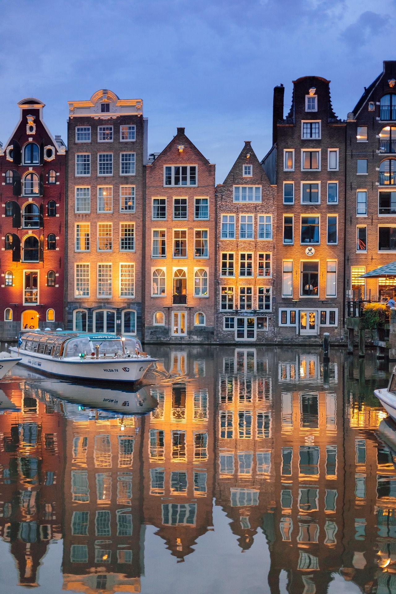 Canal-side buildings with lit windows reflected in calm water; a boat is docked, Amsterdam, dusk.
