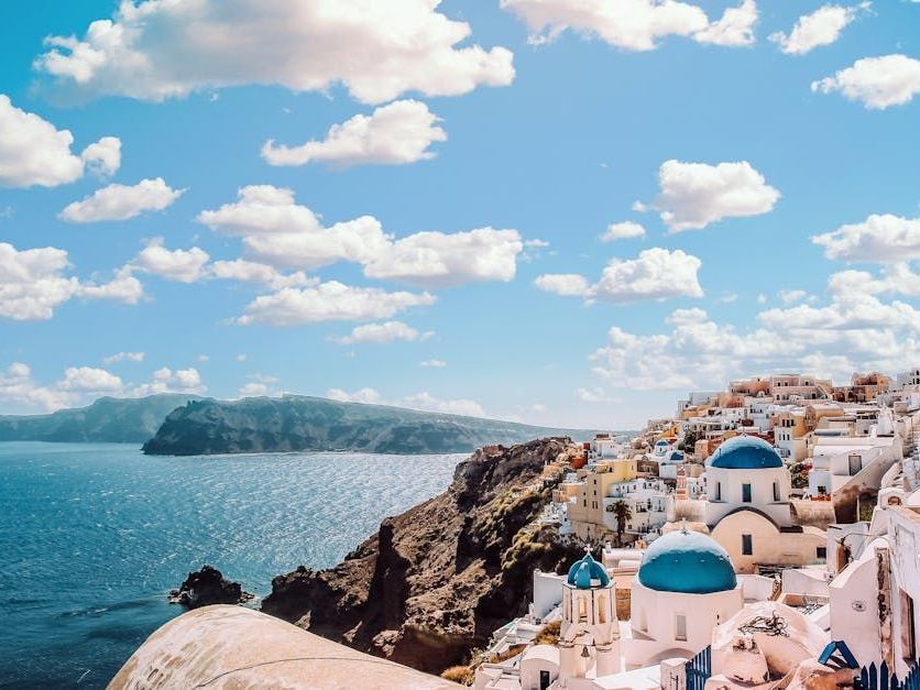 Coastal Santorini, Greece with white buildings, blue domes, and a bright blue sky with puffy clouds.
