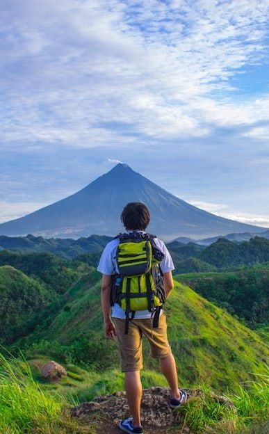 Hiker with backpack gazing at a symmetrical volcano, atop a grassy hill under a cloudy sky.