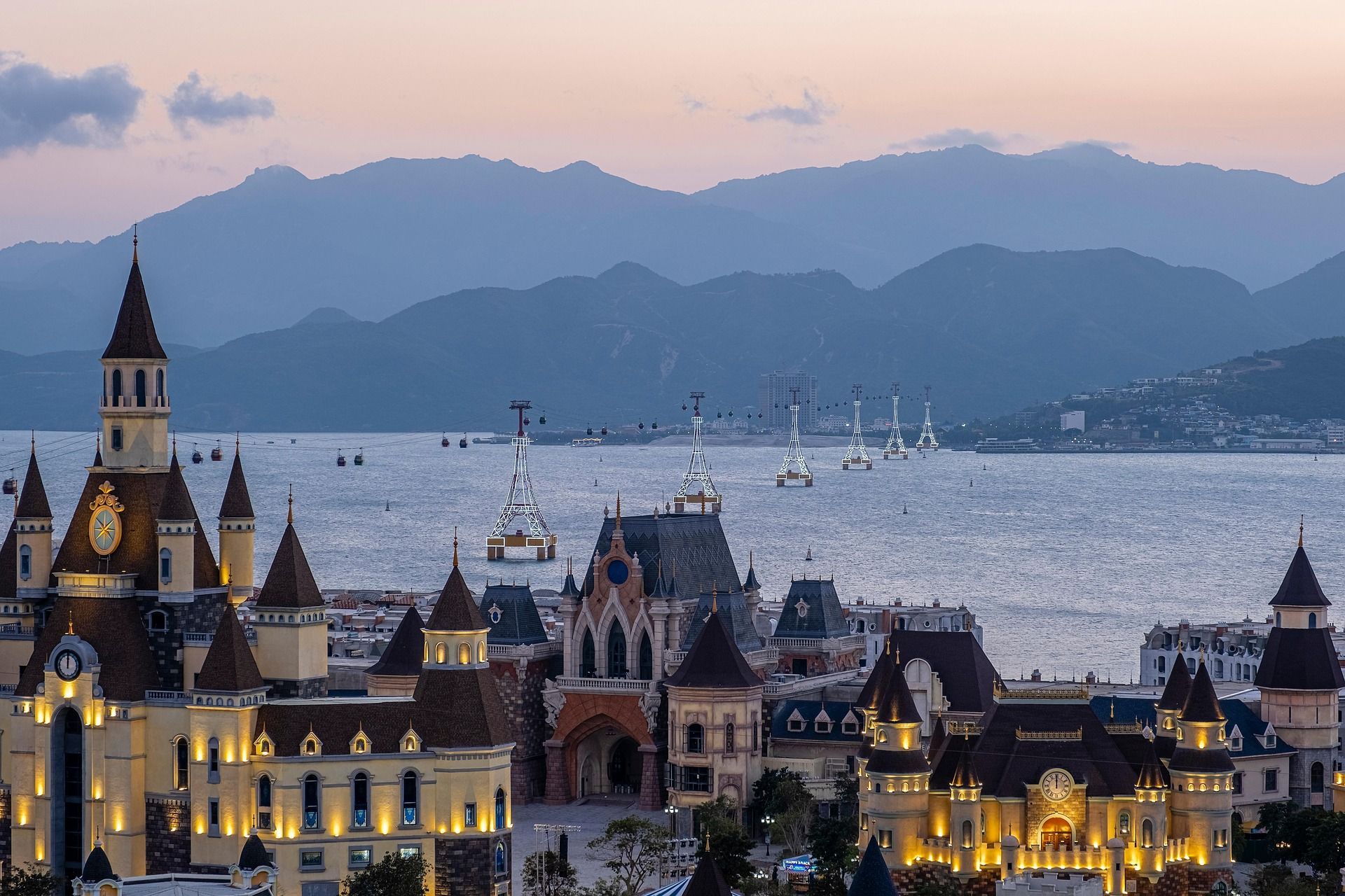 A colorful castle with spires by the sea, mountains in the distance at dusk.