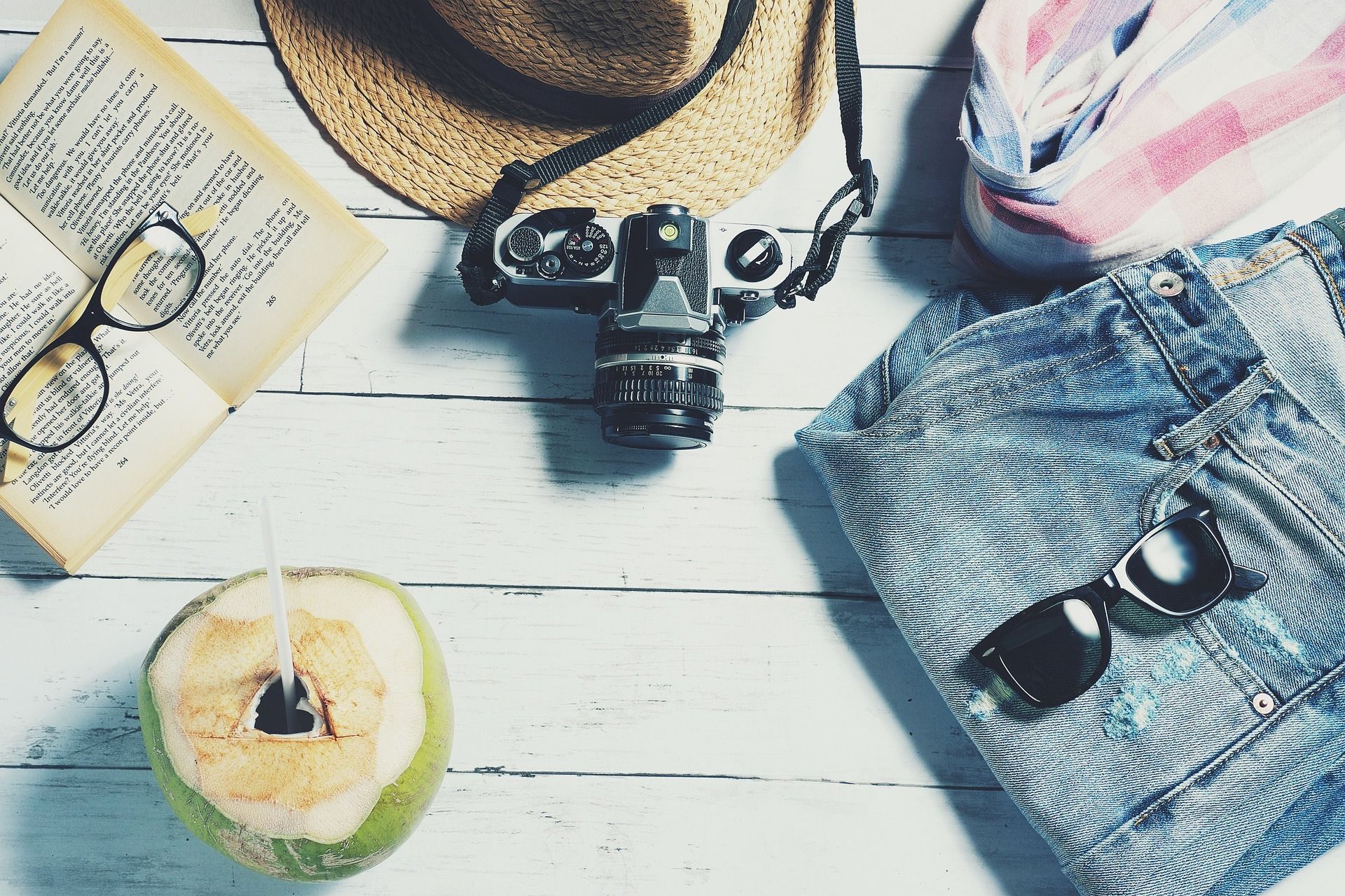 Overhead view: travel items on a white wood table. Coconut, sunglasses, camera, hat, book, and denim shorts.