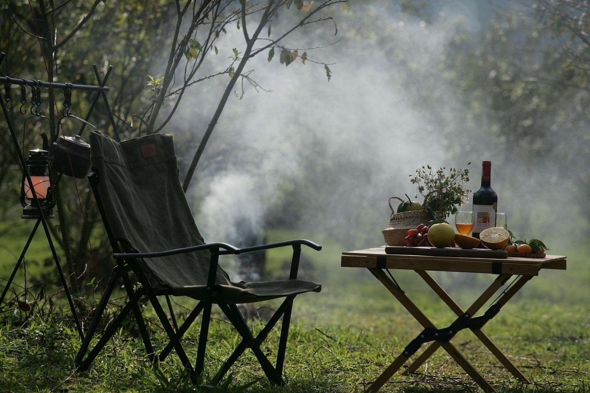 Camping scene: chair, small table with food & wine, smoke, lantern, trees.