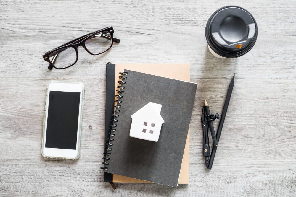 Top view of a workspace with notebooks, a house model, a phone, coffee, and tools on a wooden surface.