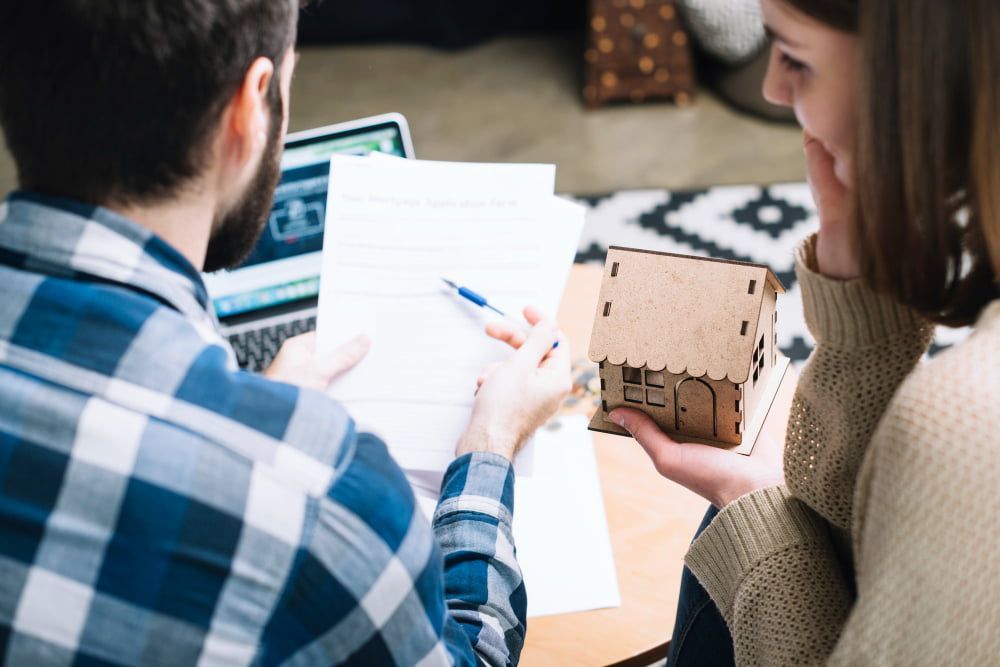 A couple reviews mortgage papers at a table; he points to documents while she holds a model house, suggesting contemplation.