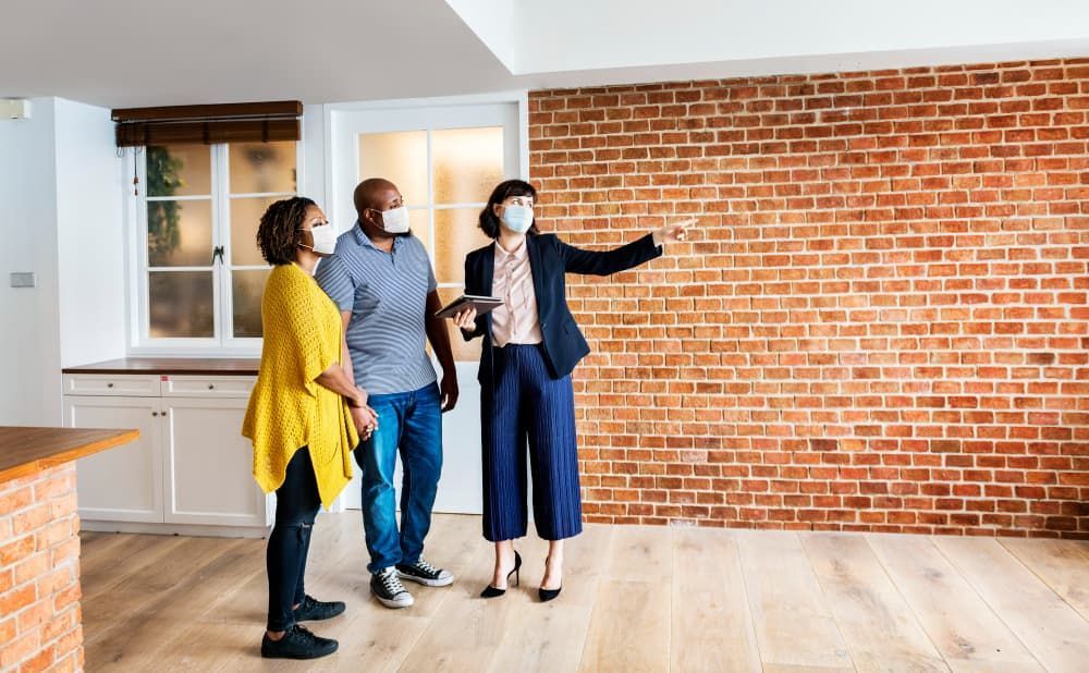 A masked real estate agent shows a brick-walled apartment to an attentive, masked couple.