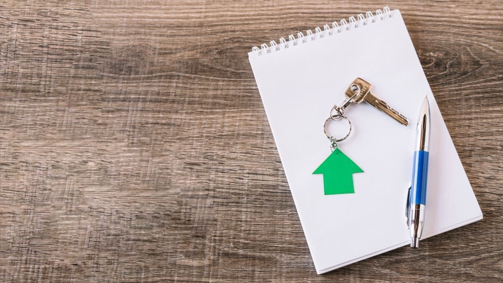 Notebook with house keychain and keys beside a pen on a wooden table, suggesting home planning or real estate.
