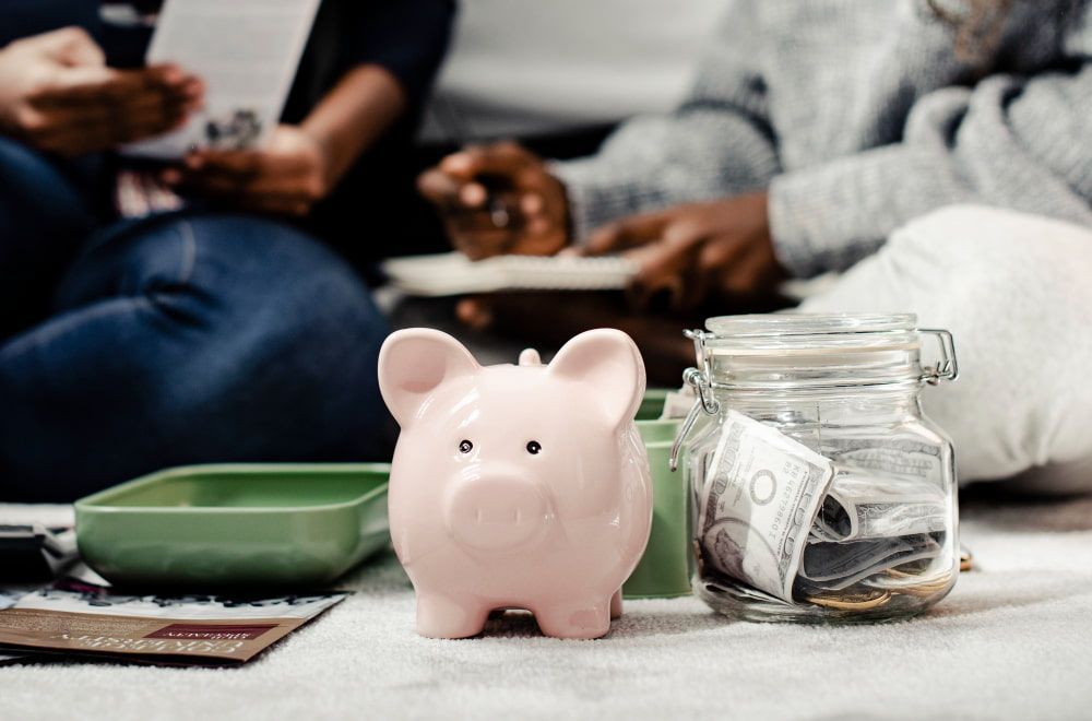 A pink piggy bank and cash-filled jar sit on a carpet as two people focus on financial planning in the background.