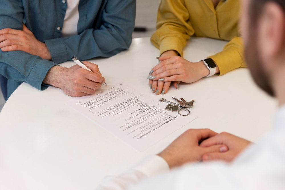 A person in a blue shirt signs a document on a white table, with keys nearby.