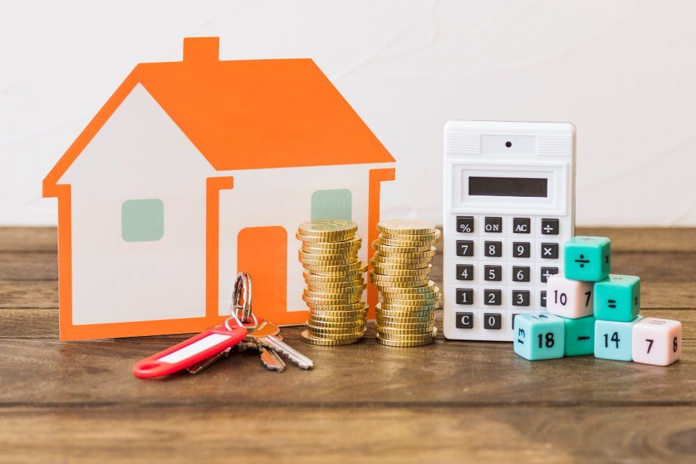 A paper house, gold coins, keys, a calculator, and colorful dice on wood symbolize real estate and finances.