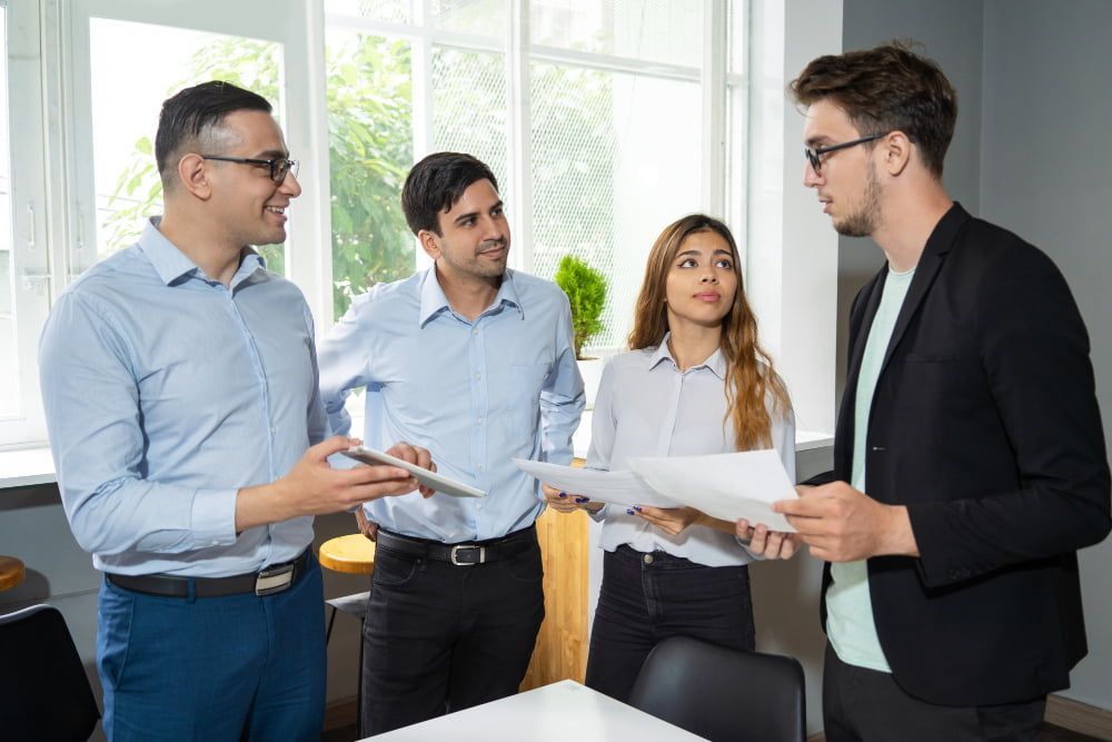 Four professionals in business casual discuss in a bright office, holding papers and a tablet, looking focused.