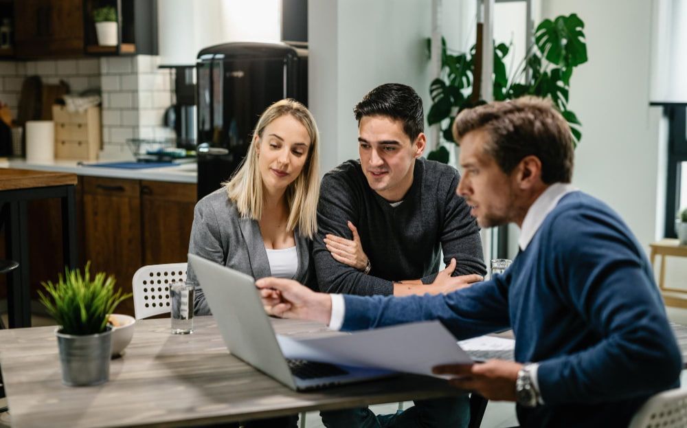 A couple attentively listens to a professional man pointing at a laptop in a modern office.