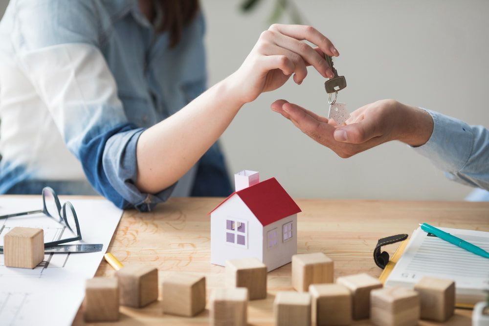 Hands exchange house keys over a table with plans and a paper house, symbolizing a hopeful, collaborative home purchase.