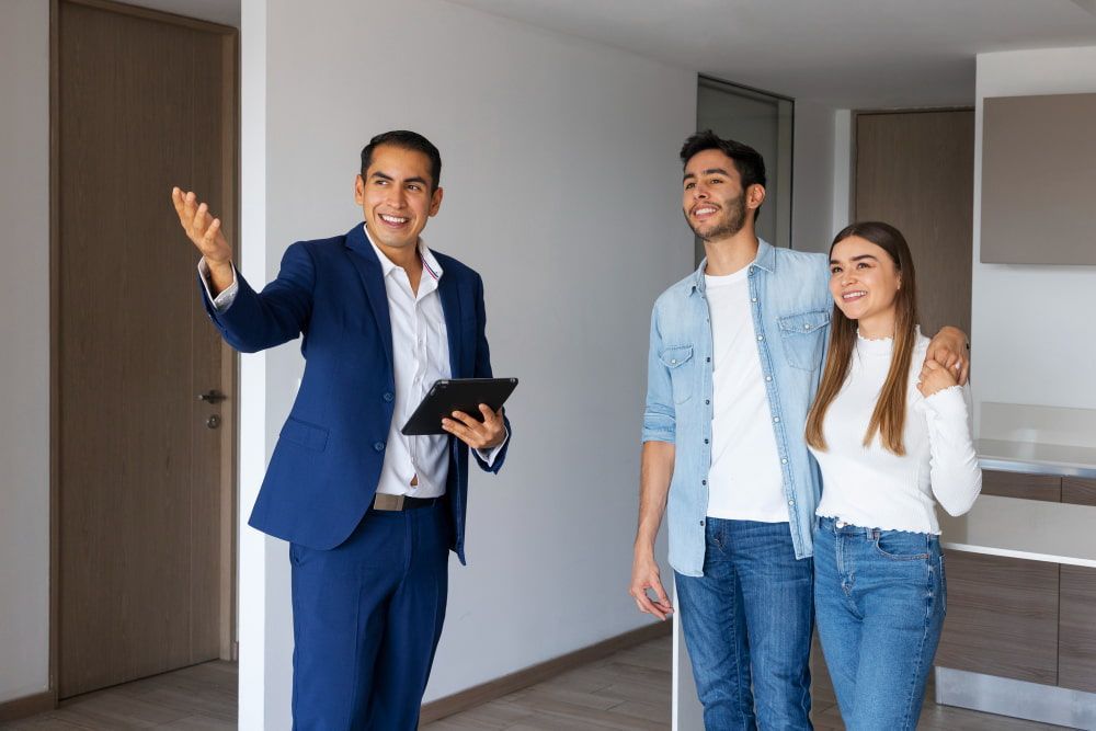 A real estate agent in a suit shows a smiling couple a modern, unfurnished room.