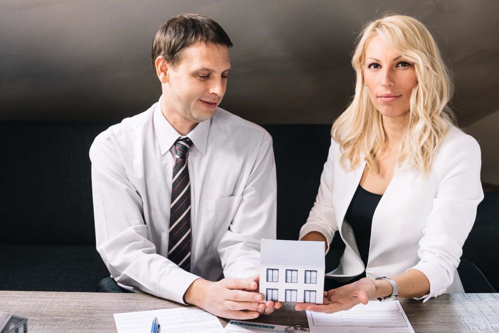 Two people hold a house model over papers and a pen at a table, creating a professional atmosphere.
