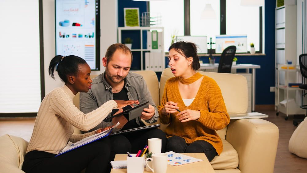 Three people sit in a modern office discussing. One shows a tablet, engaging the others, who appear focused.