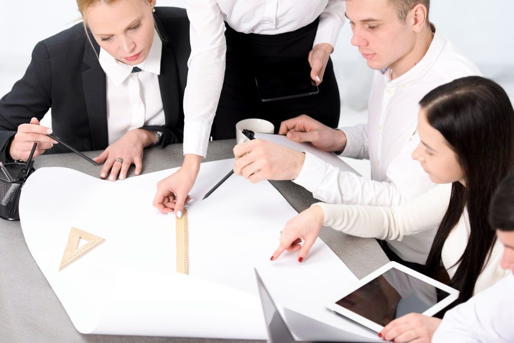 Group in formal attire collaborates with paper, rulers, and tablet, showing teamwork and planning.