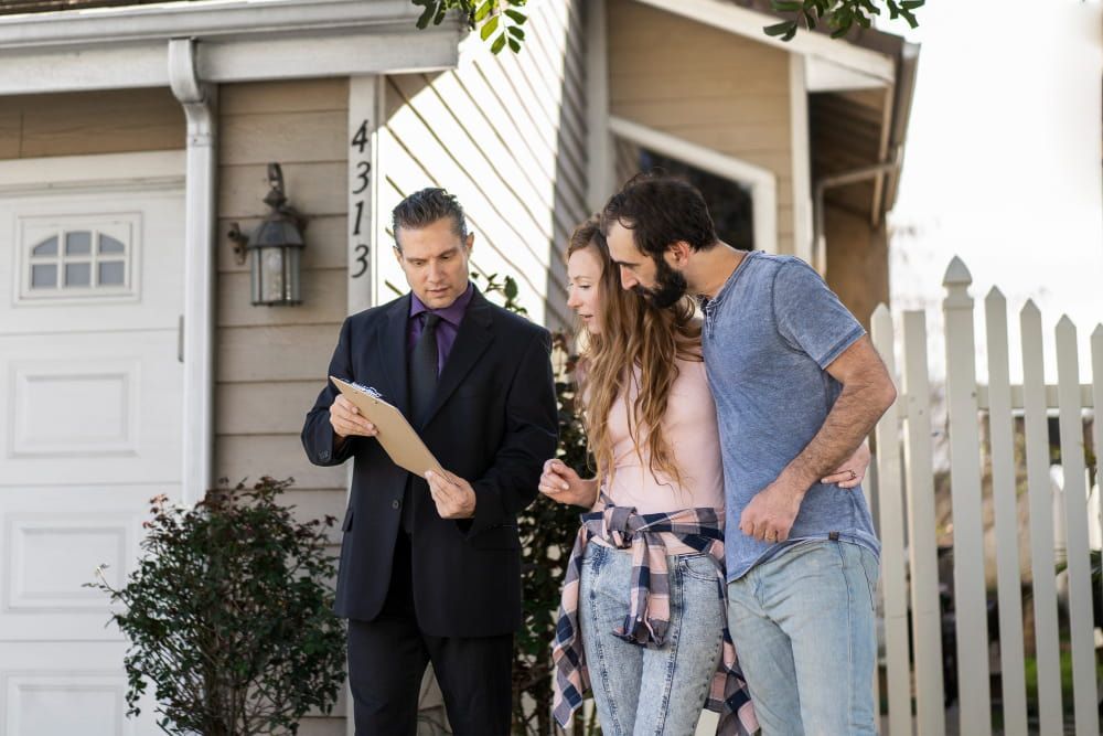 A suited real estate agent discusses a document with a smiling couple outside a beige house with a white fence.