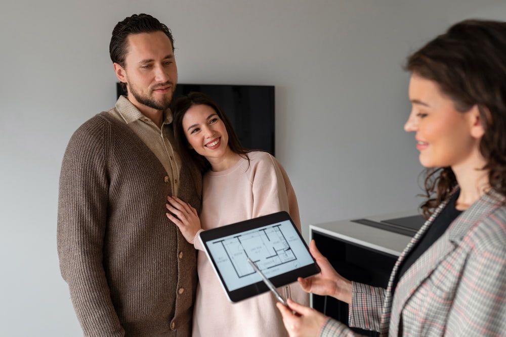 A smiling couple looks at each other as a real estate agent shows them a floor plan on a tablet.