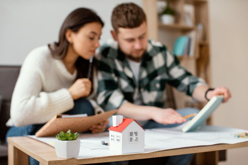 A couple sits closely, focused on house plans at a table with a small model house and a succulent.