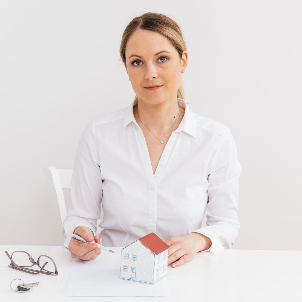 A woman in a white shirt holds a pen and house model at a table, suggesting real estate planning; glasses and keys nearby.