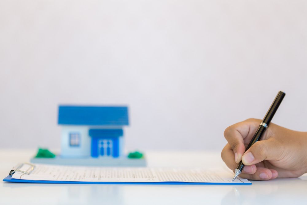 A hand signs a document on a clipboard; a blurred model house with a blue roof in the background suggests a mortgage signing.