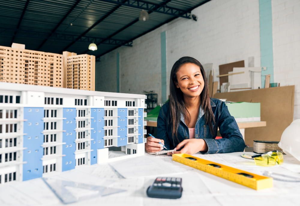 A smiling woman in denim works with models and blueprints in a bright workshop, showing creativity and focus.