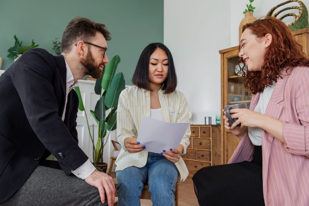 Three people sit in a cozy, green-walled room with plants as a woman reads a paper and the others listen attentively.