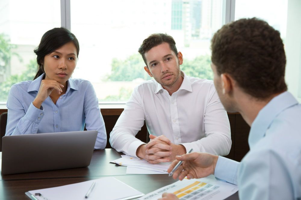Three pros discuss at a table with charts and laptop; one gestures, others listen, showing focus.


