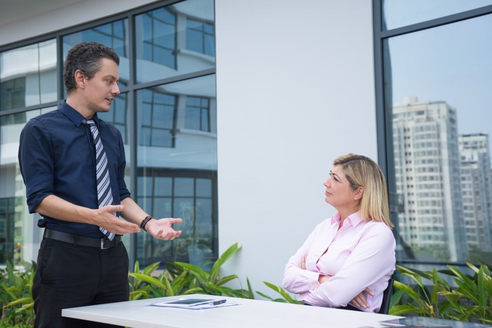 A man in a tie gestures while speaking to a thoughtful woman in pink outside a modern building.