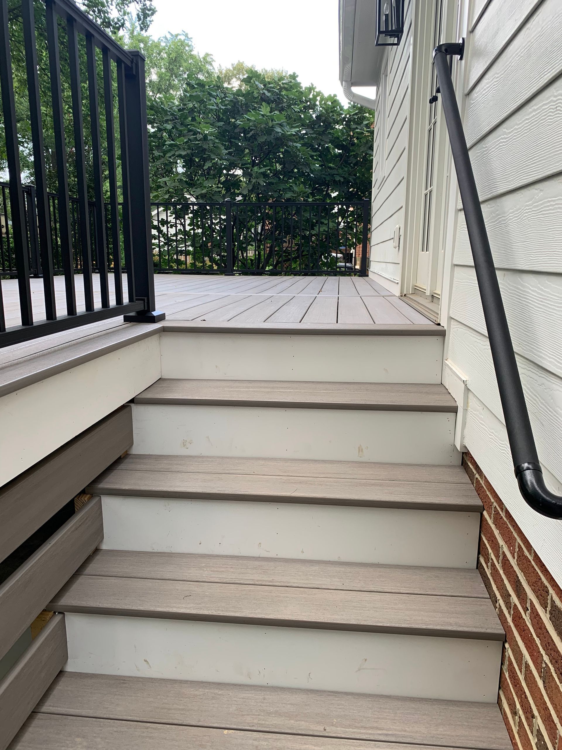 Steps leading up to a wooden deck with black railing and a black handrail on the adjacent house.