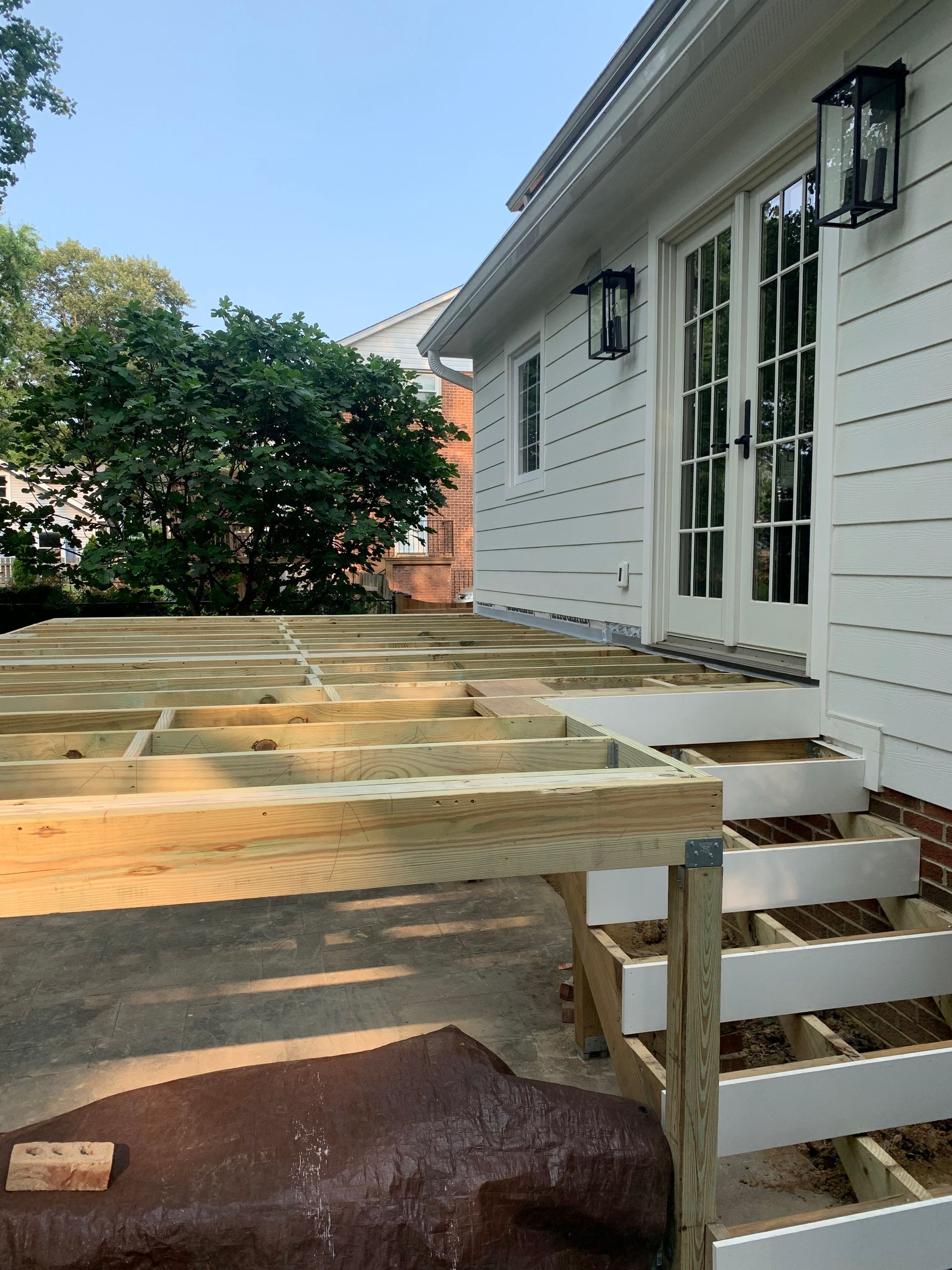 Unfinished wooden deck with stairs next to a white house with double doors and black sconces.