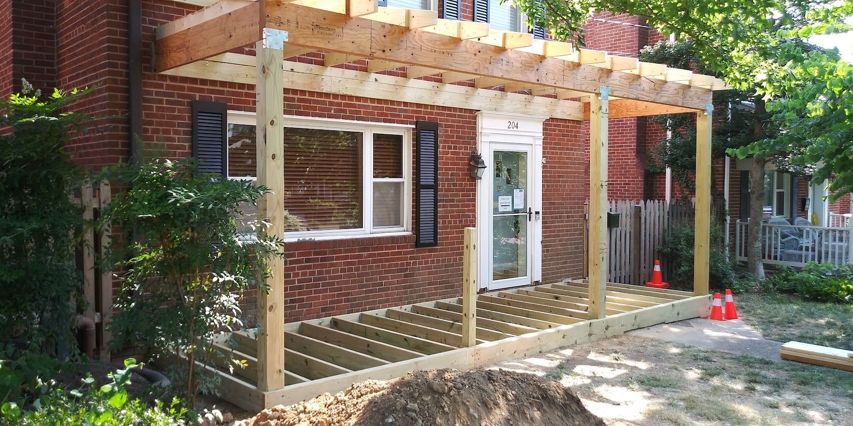 A porch under construction. New wooden deck and pergola frame attached to a brick house.