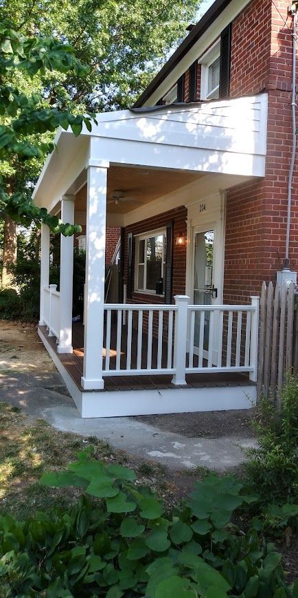 White porch with railings, brown deck, and white columns attached to a red brick house.