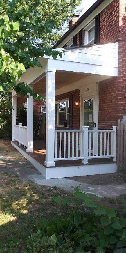 White porch with railing and columns attached to a brick house. Trees and greenery surround the house.