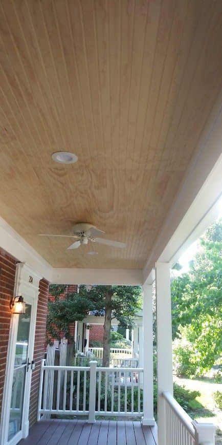 Porch ceiling with wood paneling, light fixture, and ceiling fan. Exterior view with trees, railing, and brick building.