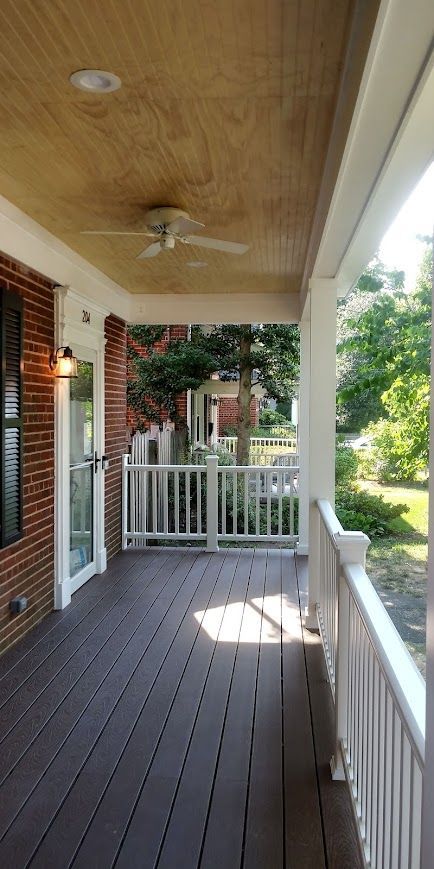 A covered porch with wood-look flooring and white railings. A ceiling fan hangs above. Brick wall on the left.