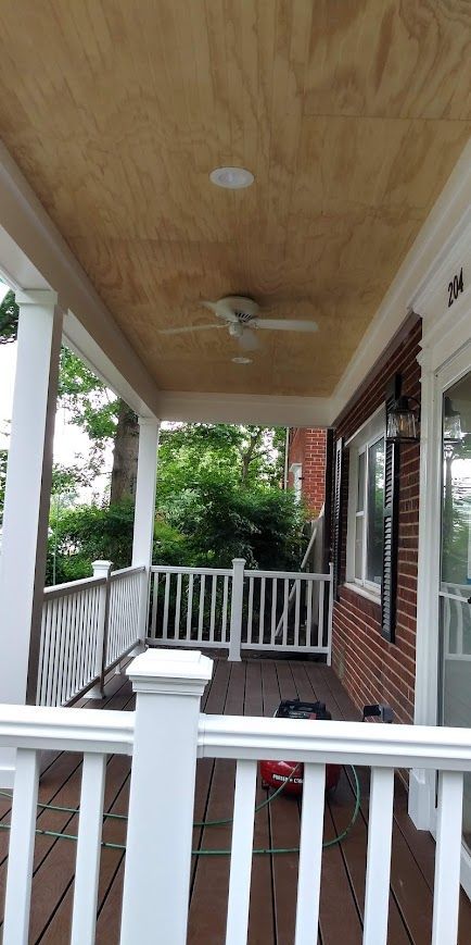 A covered porch with white railings and a wooden ceiling, a ceiling fan, and a view of trees.