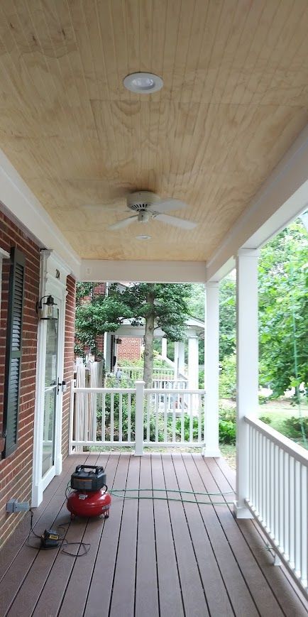 Porch with wood ceiling, fan, and red air compressor. White railings, door, and columns. Green trees in background.