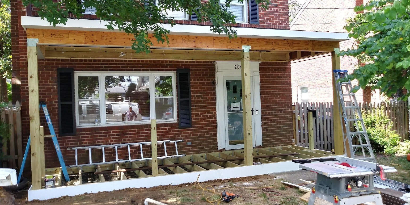 Porch construction underway on a brick house. Wood beams and posts are being erected. A saw and ladder are present.