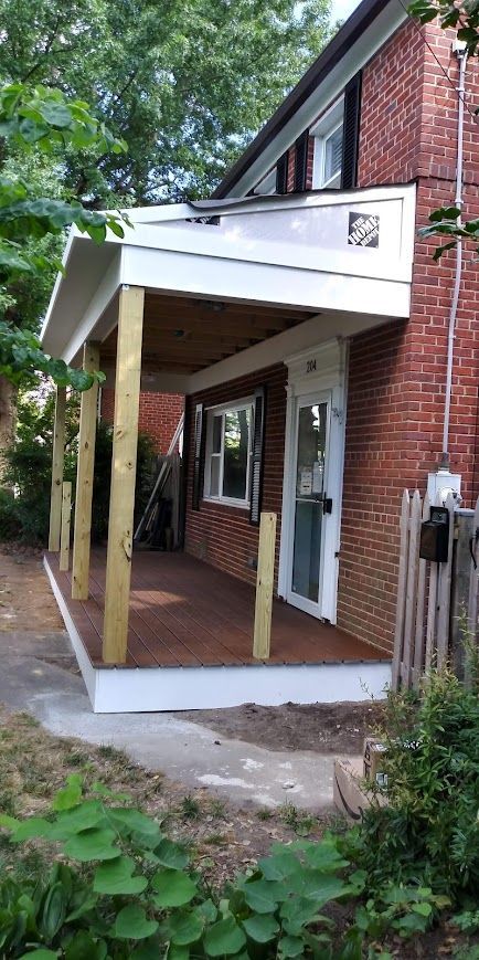Porch with white trim and wooden posts attached to a red brick house with a door and window.