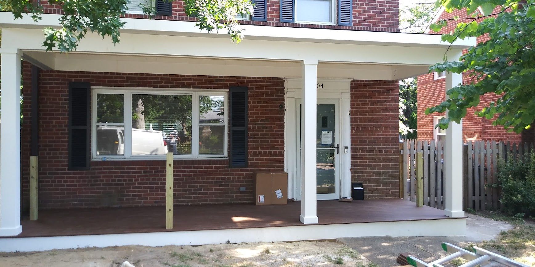 A brick house with a porch. The porch has white columns and a red-brown floor. Black shutters flank a window.