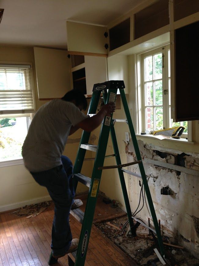 Person on a step ladder in a kitchen, reaching up to cabinetry during renovation.