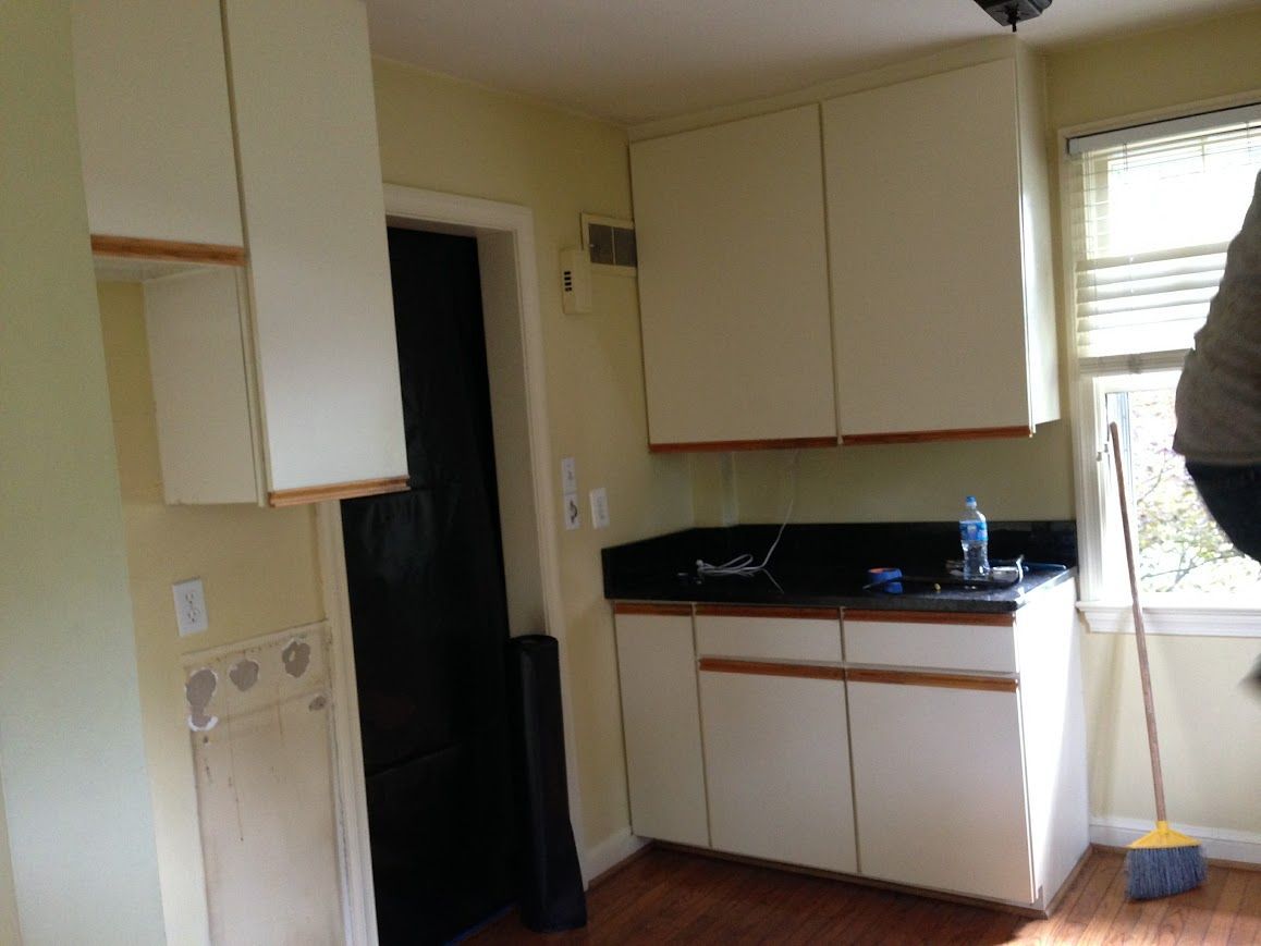Kitchen with white cabinets, dark countertop, and wooden floor. A doorway is in the center. A broom leans against the wall.