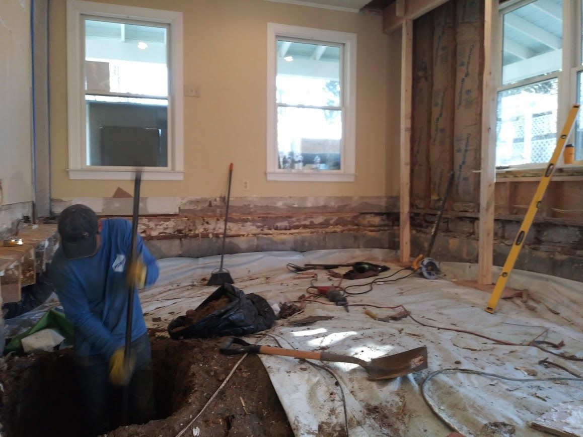 Person digging in a hole inside a room under construction. Dirt, tools, and plastic sheeting are visible.