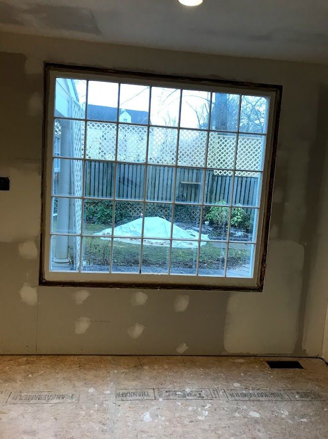 Large, multi-paned window looking out onto a backyard. Interior construction with exposed drywall and flooring.