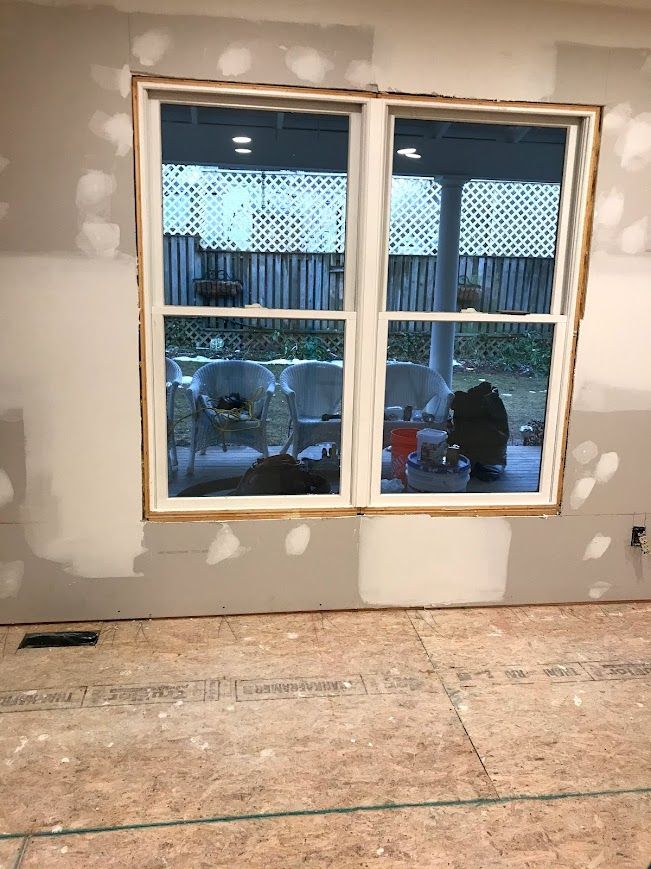 Window frame with exposed wood trim, drywall, and unfinished flooring. Window overlooks backyard.