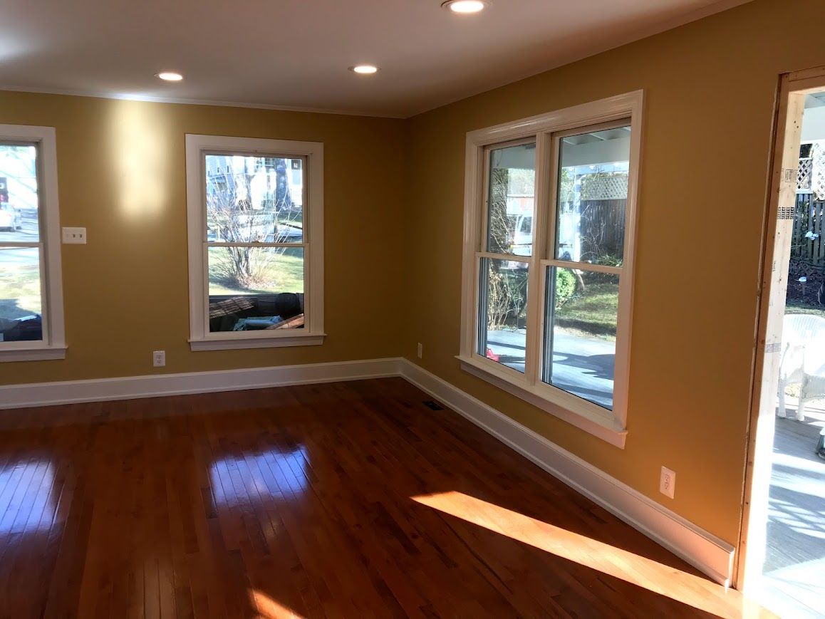 Empty room with hardwood floors, tan walls, white trim, and sunlight streaming through windows.